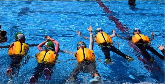 この写真は、小学生が着衣泳講習でライフジャケットを着用し、水面で仰向けになって、浮いて助けを待つ練習をしている様子です。